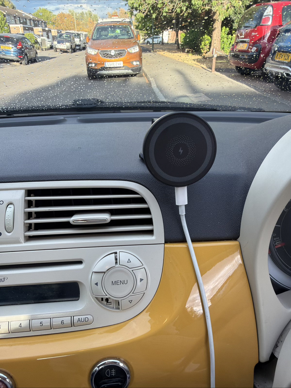 A black circular car dashboard mount holding a white wired device, installed on the dashboard of a car with a yellow and white interior. Through the windshield, a street scene with other cars is visible.